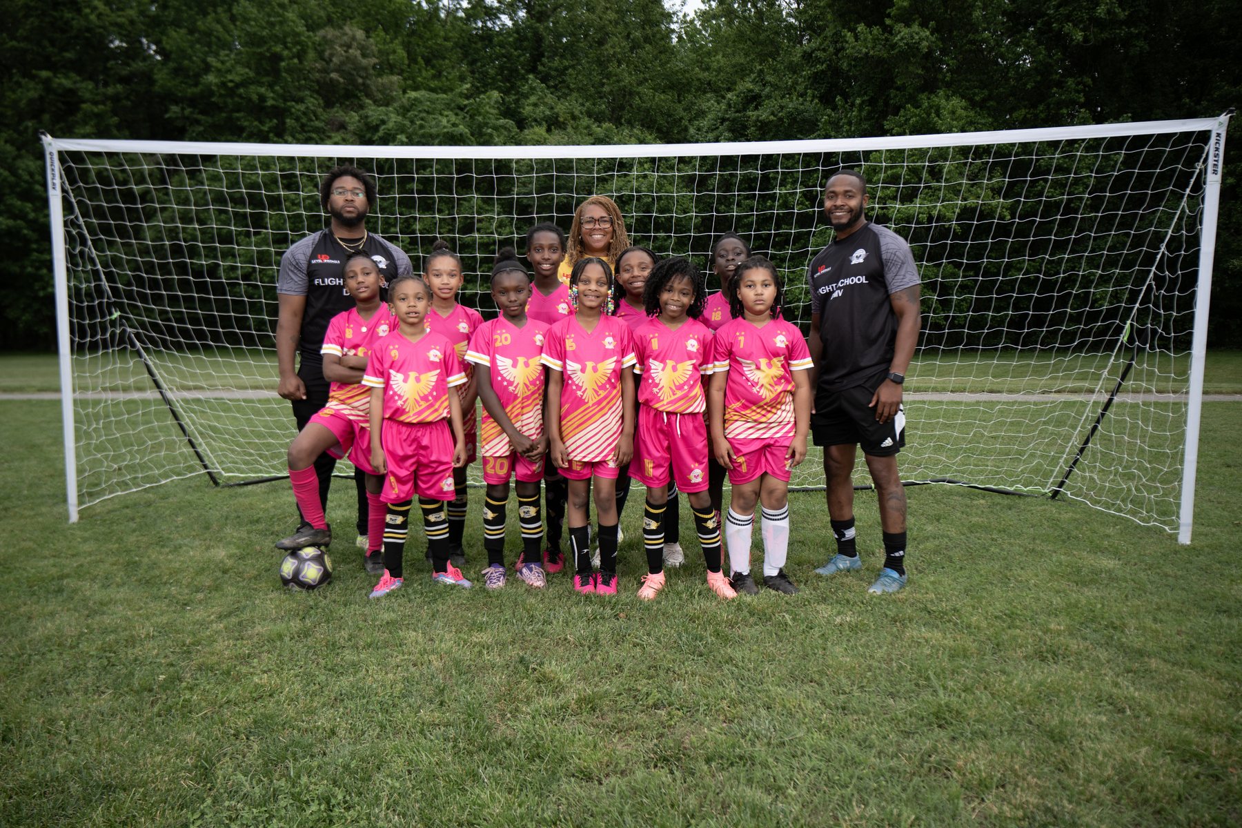 The Flight School Project girls team in pink with their coaches in front of the goal